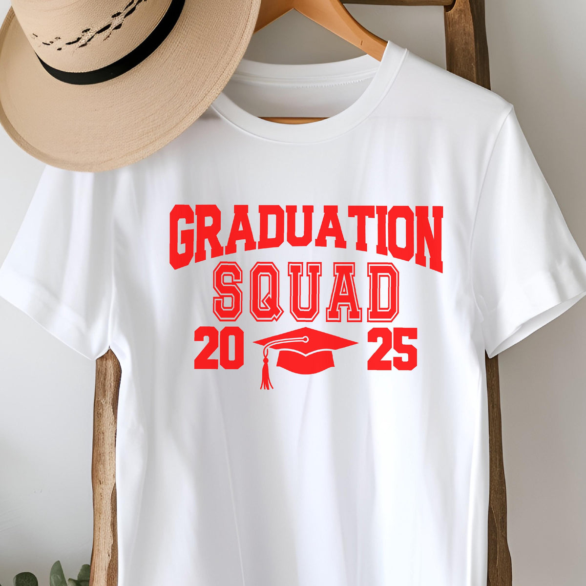 May include: White t-shirt with the words "GRADUATION SQUAD 2025" in red block letters. A red graduation cap graphic is between the numbers 20 and 25. A tan hat hangs above the shirt.