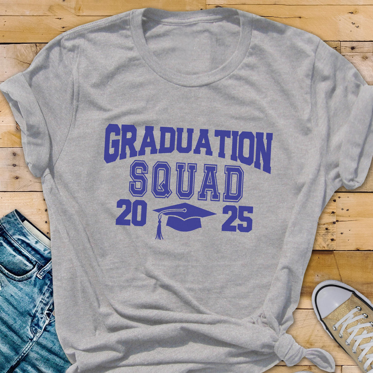 May include: A gray t-shirt with the words "GRADUATION SQUAD 2025" in blue, along with a graduation cap graphic. The shirt is made of a soft material and is suitable for casual wear. The shirt is displayed on a wooden surface.