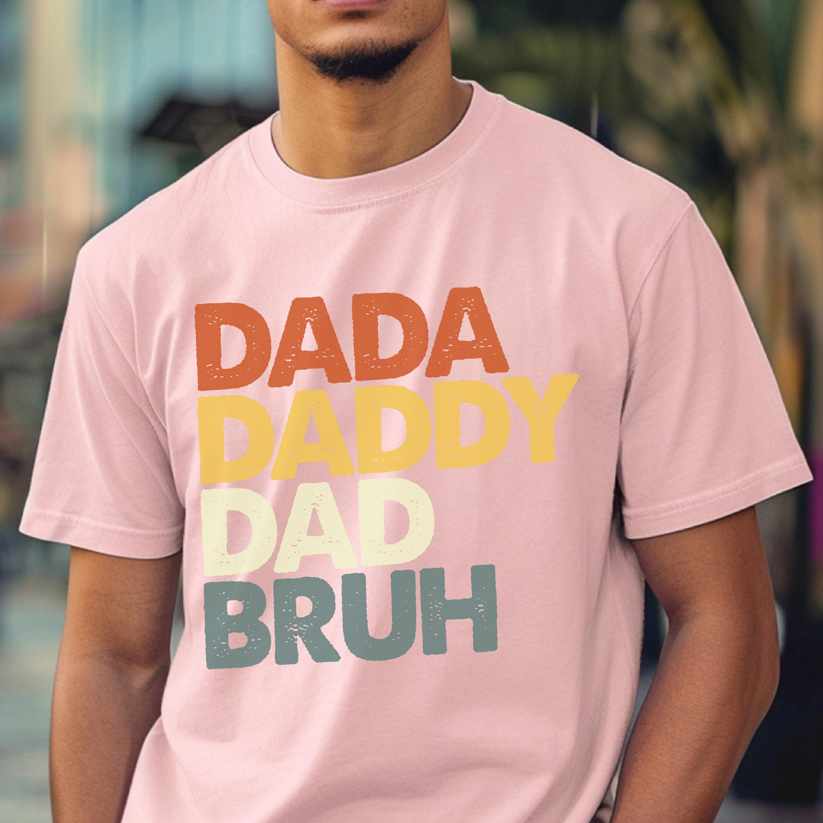 May include: A light pink t-shirt with the words "DADA DADDY DAD BRUH" in orange, yellow, light green, and blue lettering. The shirt has a crew neck and short sleeves.