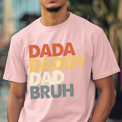 May include: A light pink t-shirt with the words "DADA DADDY DAD BRUH" in orange, yellow, light green, and blue lettering. The shirt has a crew neck and short sleeves.