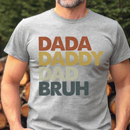 May include: A gray t-shirt with the words "DADA DADDY DAD BRUH" in a stacked, retro-style design. The text is in shades of brown, yellow, and green. The shirt is short-sleeved.
