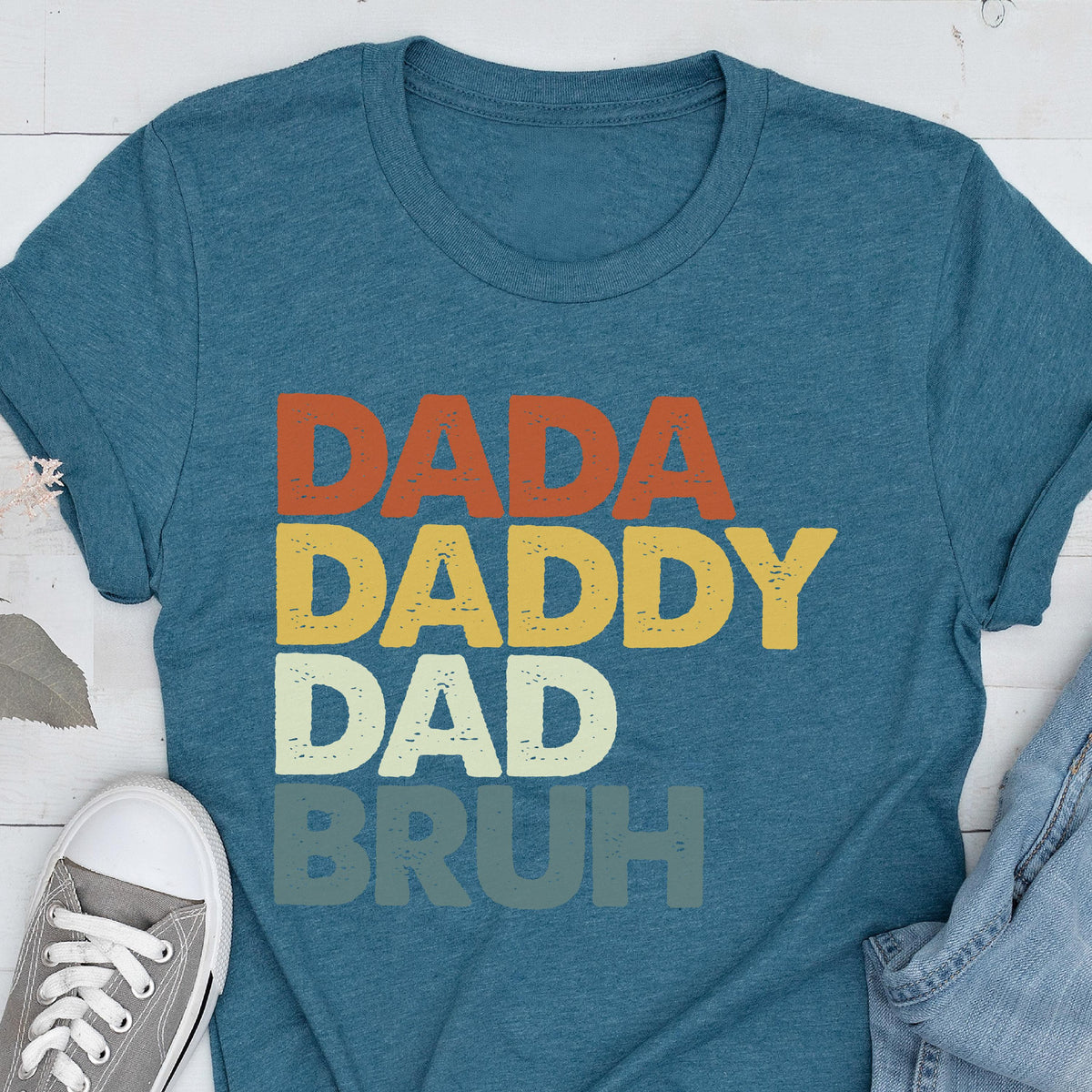 May include: Teal t-shirt with the words "DADA", "DADDY", "DAD", and "BRUH" in orange, yellow, white, and teal lettering. The shirt is made of a soft material and is suitable for casual wear.