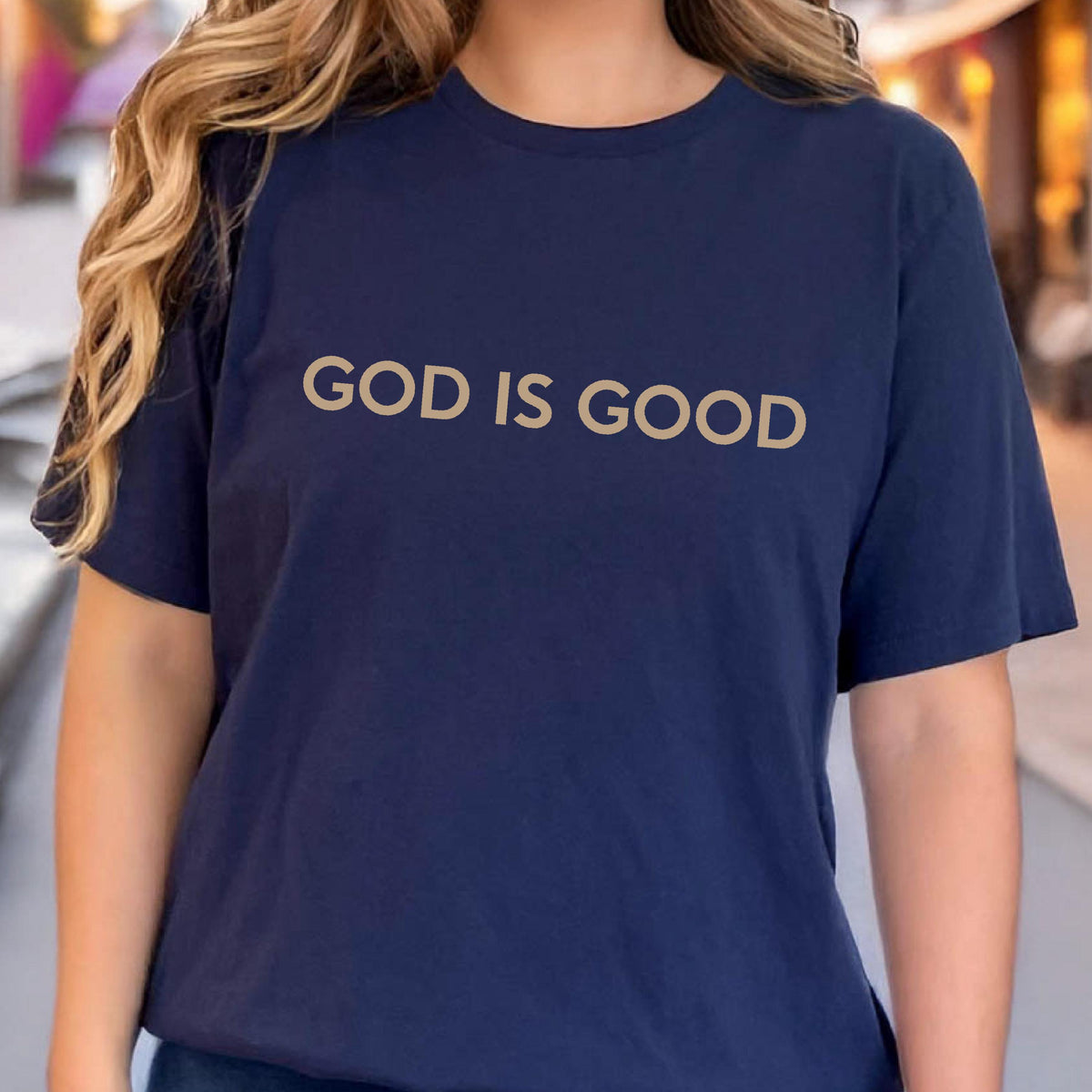 May include: Navy blue t-shirt with the text "GOD IS GOOD" in a gold color. The shirt has short sleeves and a crew neck. The image shows the upper body of a person wearing the shirt.