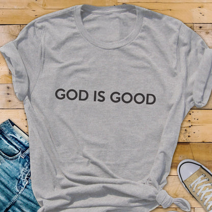 May include: A light gray t-shirt with the text "GOD IS GOOD" in black. The shirt is knotted at the bottom. A pair of blue jeans and a tan sneaker are partially visible in the background.