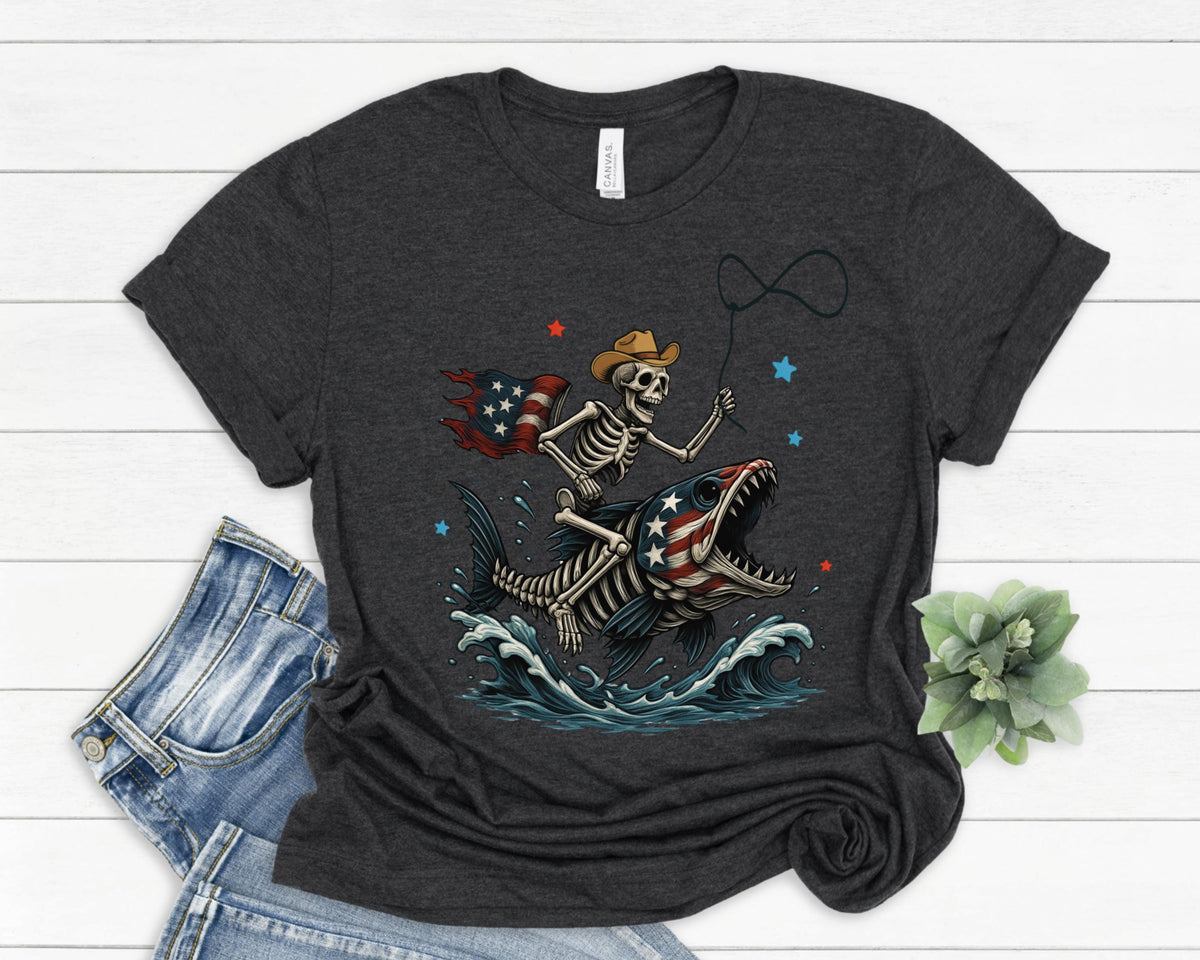 May include: Dark gray t-shirt featuring a skeleton riding a fish with an American flag design. The skeleton wears a cowboy hat and holds a lasso. The fish has an American flag design on its mouth. Blue waves and stars complete the design.