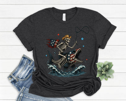 May include: Dark gray t-shirt featuring a skeleton riding a fish with an American flag design. The skeleton wears a cowboy hat and holds a lasso. The fish has an American flag design on its mouth. Blue waves and stars complete the design.