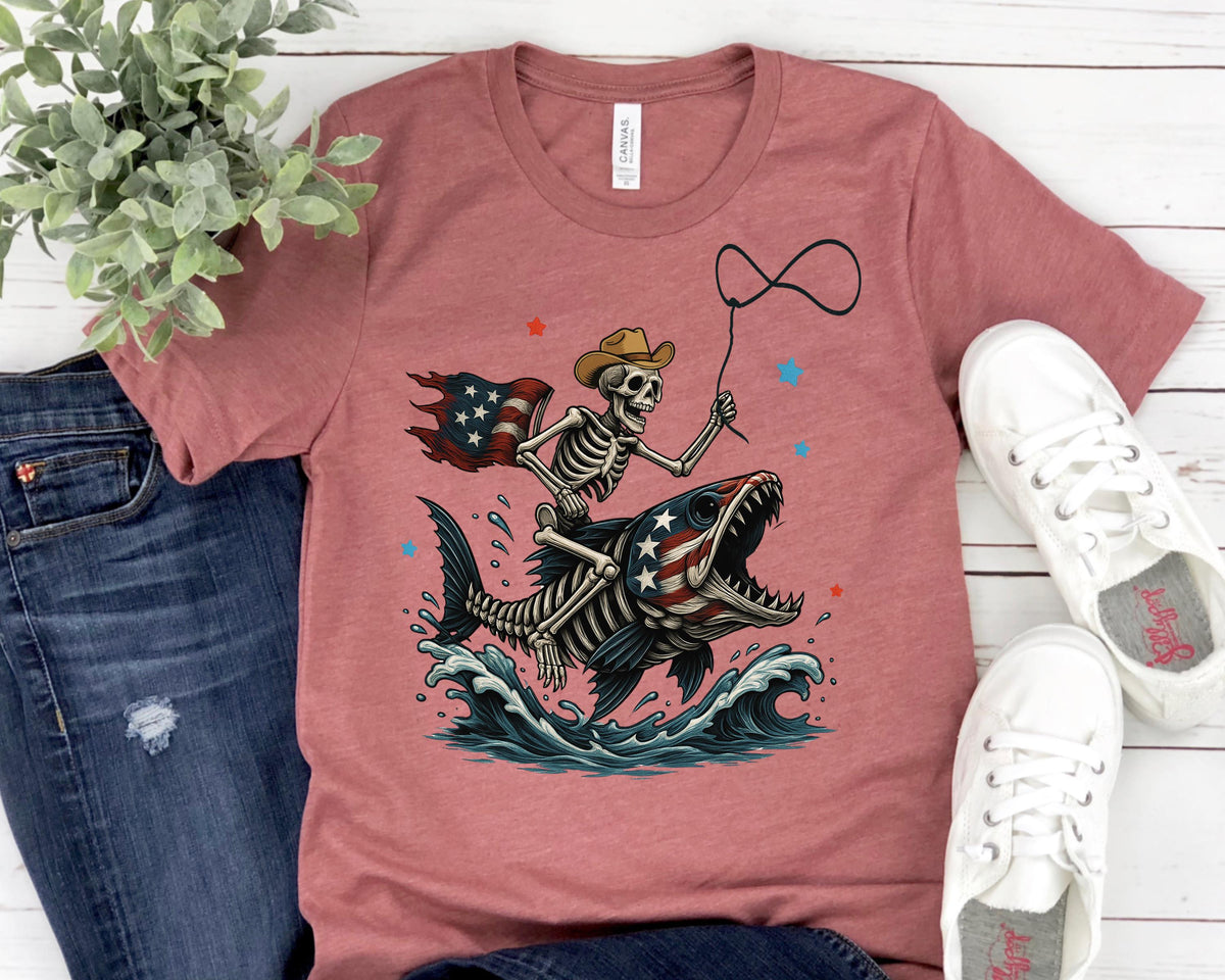 May include: A dusty rose-colored t-shirt features a skeleton riding a fish with an American flag design. The skeleton wears a cowboy hat and holds a lasso. The fish has an American flag pattern, and the waves are blue and white.