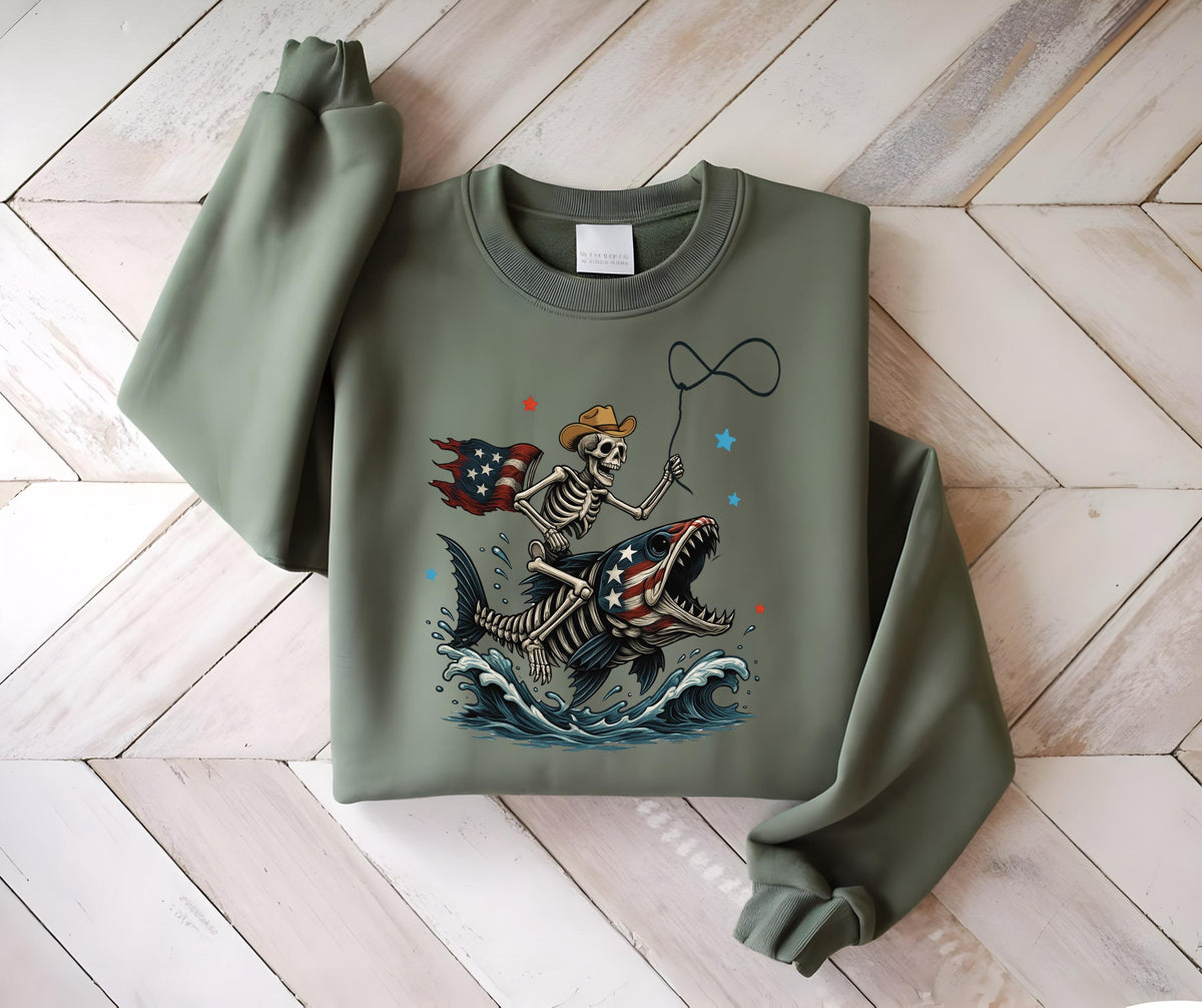 May include: Sage green sweatshirt featuring a skeleton cowboy riding a fish with an American flag design. The skeleton wears a cowboy hat and holds a lasso. The fish has an American flag pattern on its body, with waves and stars.