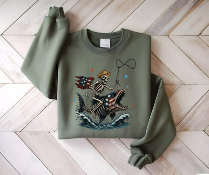 May include: Sage green sweatshirt featuring a skeleton cowboy riding a fish with an American flag design. The skeleton wears a cowboy hat and holds a lasso. The fish has an American flag pattern on its body, with waves and stars.