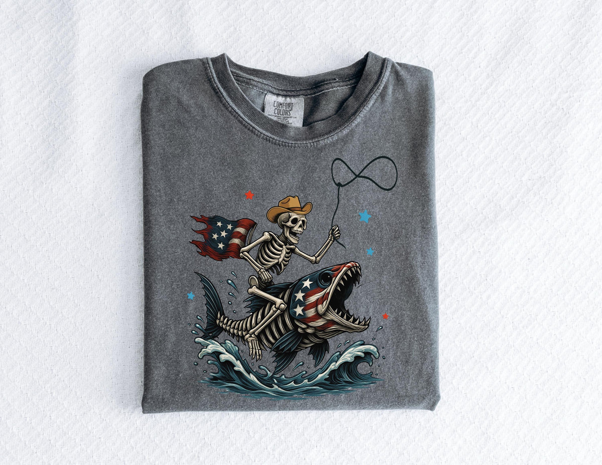 May include: A gray t-shirt featuring a skeleton cowboy riding a fish with an American flag design. The skeleton holds a lasso, and the fish has an open mouth. The design includes red and blue stars and waves.
