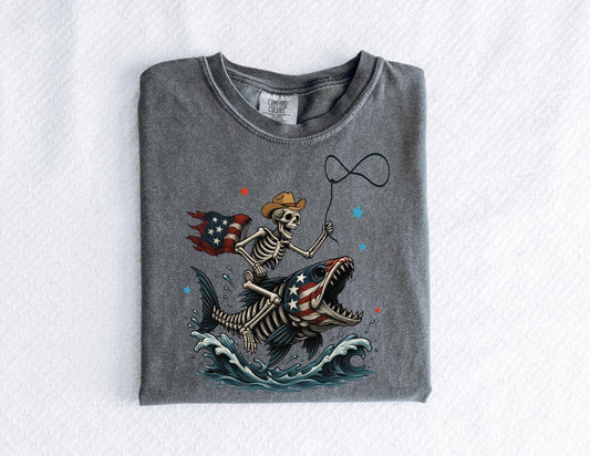 May include: A gray t-shirt featuring a skeleton cowboy riding a fish with an American flag design. The skeleton holds a lasso, and the fish has an open mouth. The design includes red and blue stars and waves.