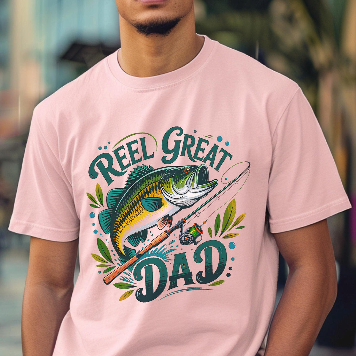 May include: A light pink t-shirt featuring a colorful graphic of a fish, fishing rod, and the words "Reel Great Dad." The design includes shades of green, blue, yellow, and brown. The shirt is a casual wear item.
