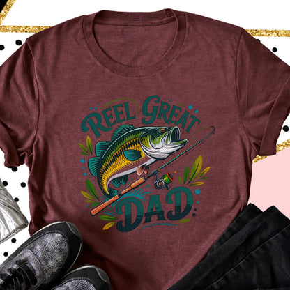 May include: Maroon t-shirt featuring a colorful graphic of a fish, fishing rod, and the words "Reel Great Dad" in teal. The fish is green, yellow, and blue. The shirt is a crew neck and has short sleeves.