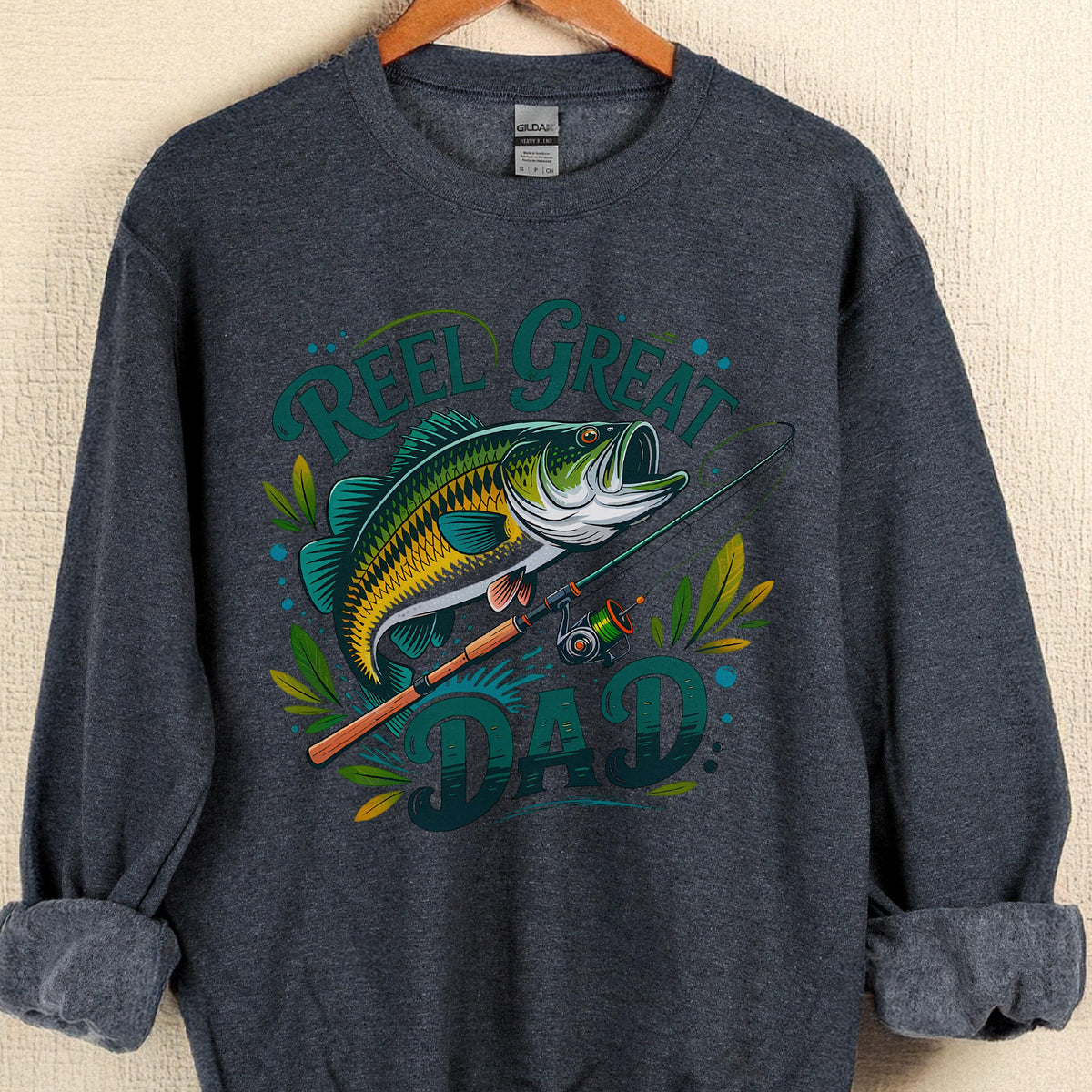May include: Dark gray sweatshirt with a graphic of a fish, fishing rod, and the words "Reel Great Dad" in teal and green. The fish is yellow, green, and white. The sweatshirt is hanging on a wooden hanger.