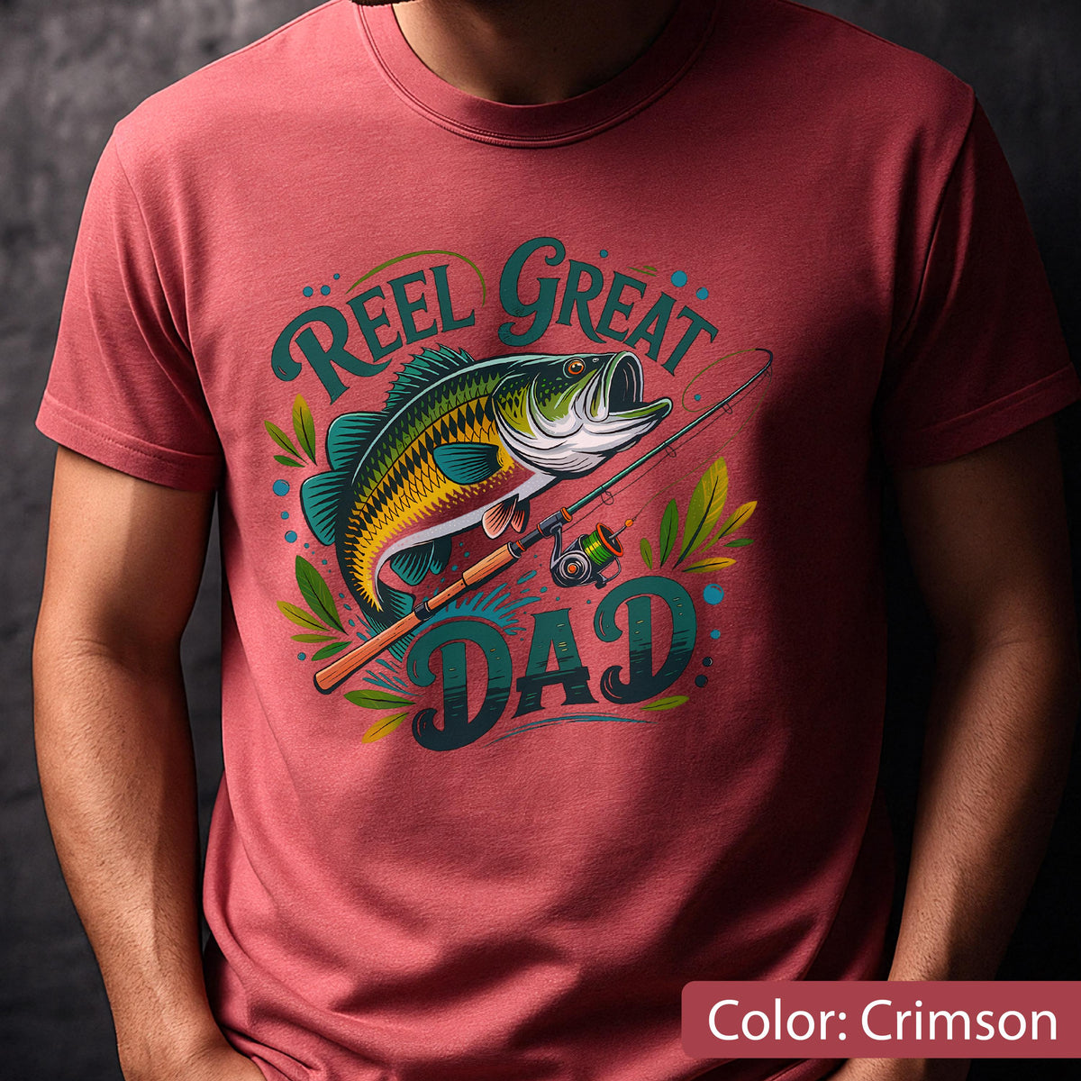 May include: Crimson t-shirt featuring a colorful graphic of a fish, fishing rod, and the words "Reel Great Dad." The design includes teal, yellow, and green accents. The shirt is a short-sleeved crew neck.
