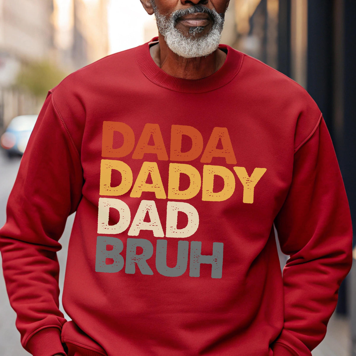 May include: A red sweatshirt with the words "DADA DADDY DAD BRUH" in a layered, retro font. The text is in shades of orange, yellow, gray, and white. The sweatshirt is a solid color and has long sleeves.