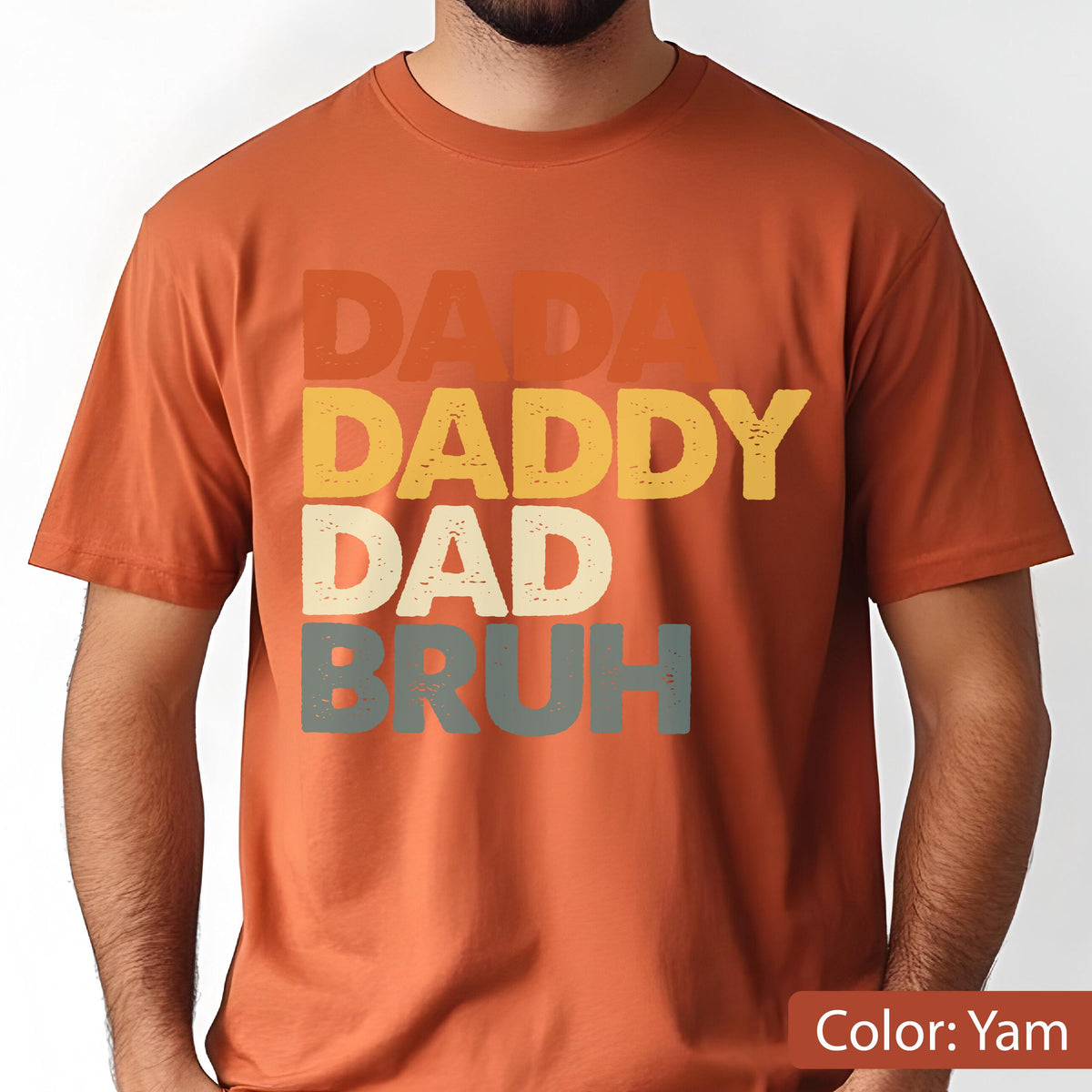 May include: A short-sleeved, yam-colored t-shirt with the words "DADA", "DADDY", "DAD", and "BRUH" stacked vertically in retro-style lettering. The text is in shades of orange, yellow, off-white, and gray.