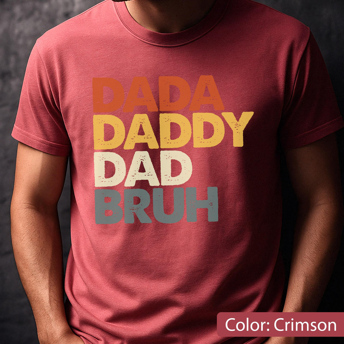 May include: Crimson-colored t-shirt with the words "DADA DADDY DAD BRUH" in a retro font. The text is in orange, yellow, off-white, and gray. The shirt is a crew neck and has short sleeves.