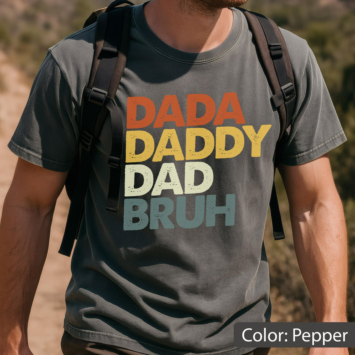 May include: Pepper-colored t-shirt with the words "DADA DADDY DAD BRUH" in orange, yellow, off-white, and teal lettering. The shirt has a vintage, distressed look. The color of the shirt is described as "Pepper".