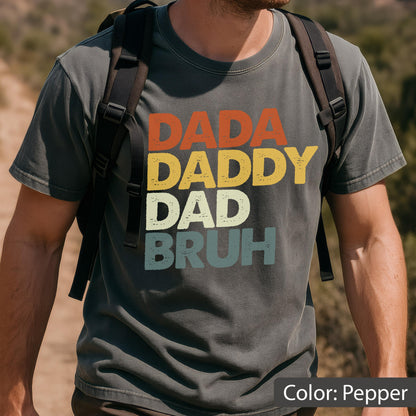 May include: Pepper-colored t-shirt with the words "DADA DADDY DAD BRUH" in orange, yellow, off-white, and teal lettering. The shirt has a vintage, distressed look. The color of the shirt is described as "Pepper".