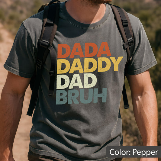 May include: Pepper-colored t-shirt with the words "DADA DADDY DAD BRUH" in orange, yellow, off-white, and teal lettering. The shirt has a vintage, distressed look. The color of the shirt is described as "Pepper".