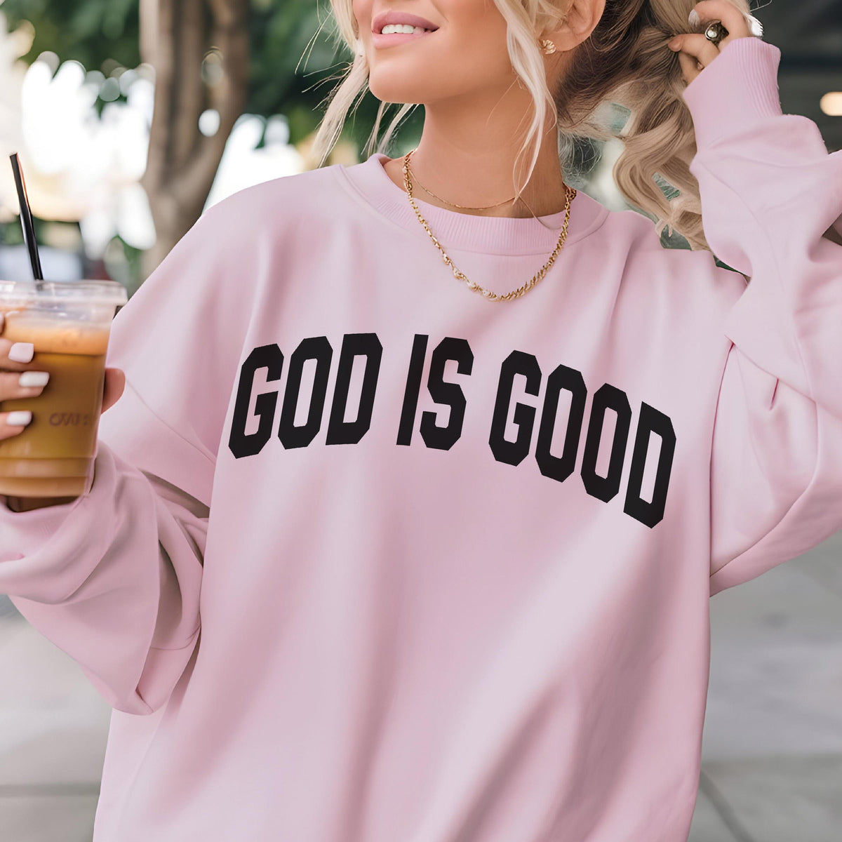 May include: A light pink sweatshirt with the text "GOD IS GOOD" in bold black letters. The sweatshirt has long sleeves and a crew neck. A gold necklace is visible.