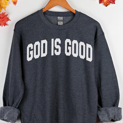 May include: A dark gray sweatshirt with the text "GOD IS GOOD" in bold white letters. The sweatshirt is displayed on a wooden hanger, with fall leaves in the background. The sweatshirt has a crew neck and long sleeves.