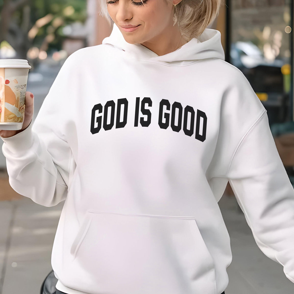 May include: White hoodie with the text "GOD IS GOOD" in bold, black letters across the chest. The hoodie has a front pocket and a drawstring hood. The person in the image is holding a coffee cup.