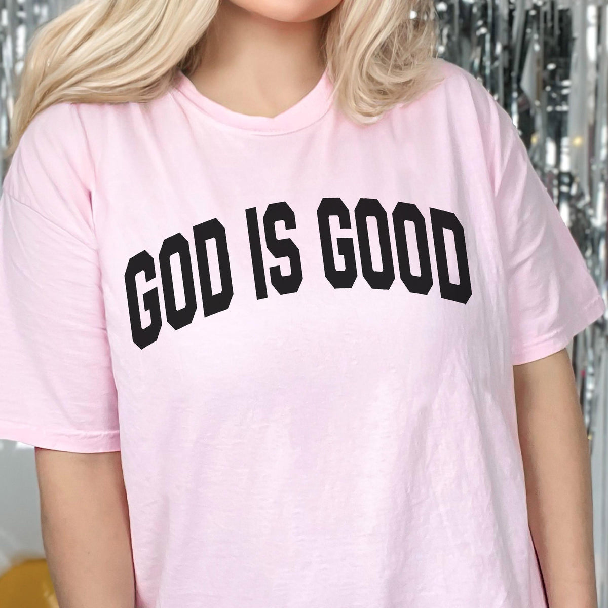 May include: A light pink t-shirt with the phrase "GOD IS GOOD" printed in bold, black letters across the chest. The shirt has short sleeves and a crew neck.