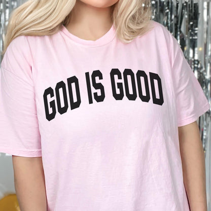 May include: A light pink t-shirt with the phrase "GOD IS GOOD" printed in bold, black letters across the chest. The shirt has short sleeves and a crew neck.
