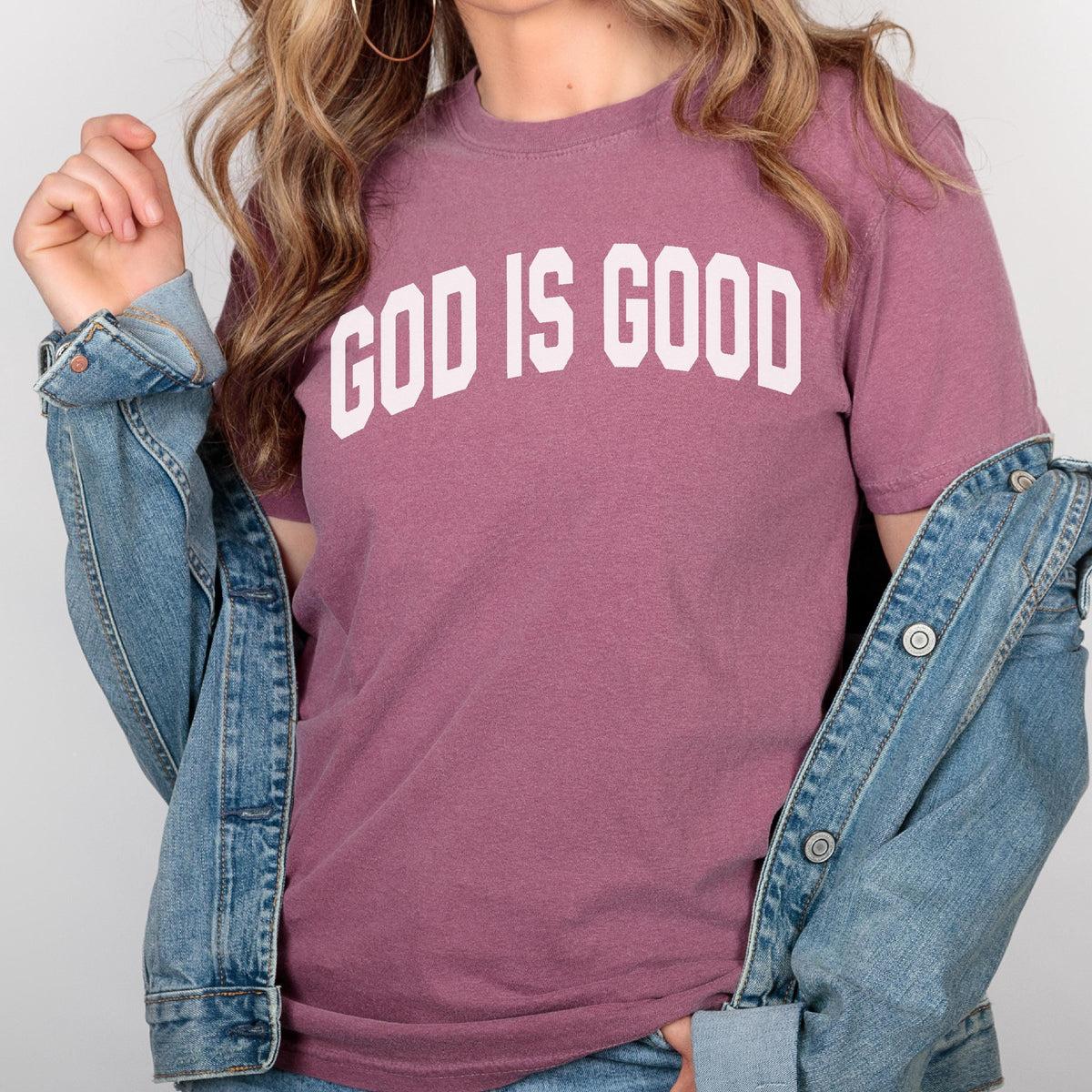 May include: A mauve-colored t-shirt with the text "GOD IS GOOD" in bold, white, block letters. A denim jacket is draped over the shoulders. The shirt has short sleeves and a crew neck.