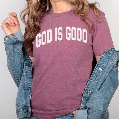 May include: A mauve-colored t-shirt with the text "GOD IS GOOD" in bold, white, block letters. A denim jacket is draped over the shoulders. The shirt has short sleeves and a crew neck.