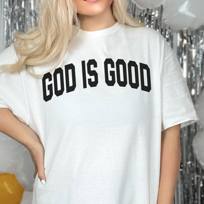 May include: White t-shirt with the text "GOD IS GOOD" in bold, black, block letters. The shirt has a crew neck and short sleeves. The background includes silver tinsel and white balloons.
