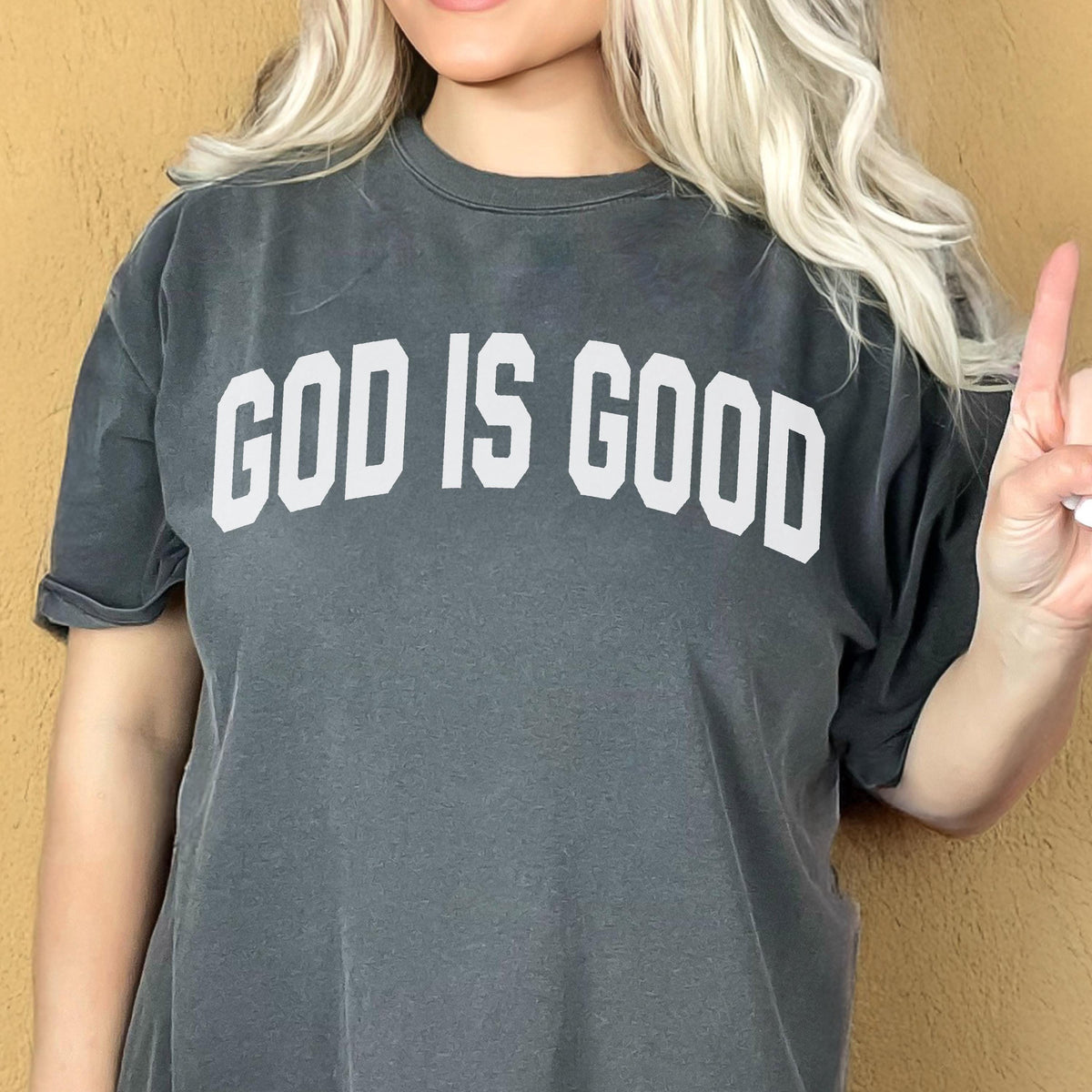 May include: Dark gray t-shirt with the text "GOD IS GOOD" in white block letters. The shirt has short sleeves and a slightly faded appearance. The background is a solid, warm, neutral color.