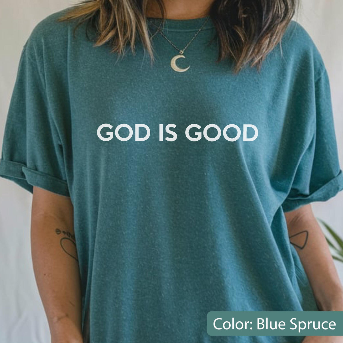 May include: Blue spruce-colored t-shirt with the text "GOD IS GOOD" in white block letters. The shirt has rolled-up sleeves and a relaxed fit. The color is described as blue spruce.