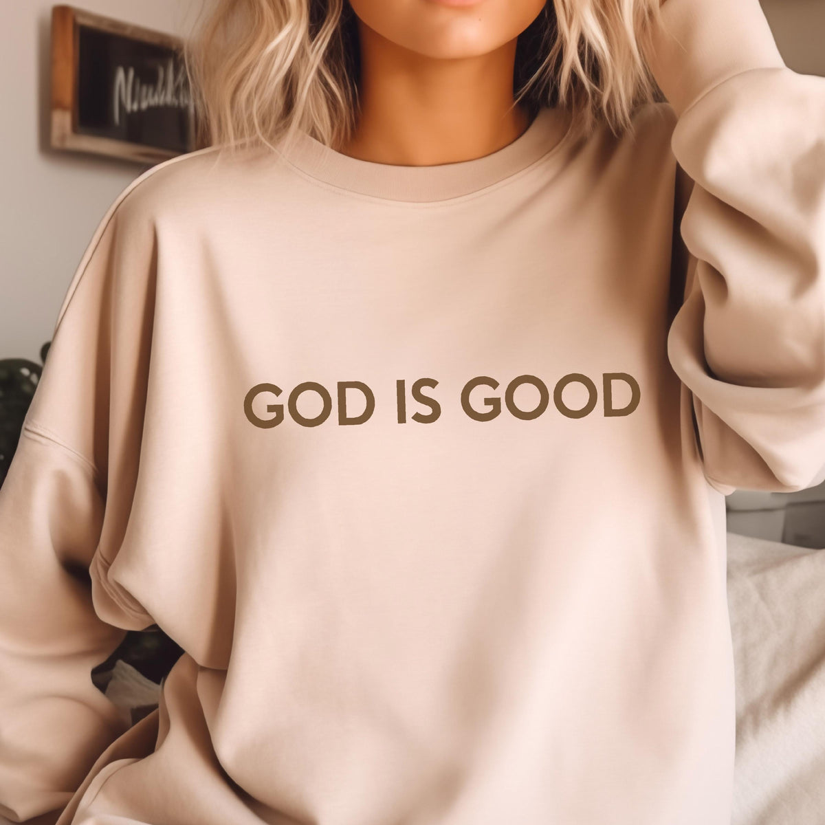 May include: A beige sweatshirt with the phrase "GOD IS GOOD" printed in brown letters. The sweatshirt is a neutral color and has long sleeves. The image is a close-up of the upper body.