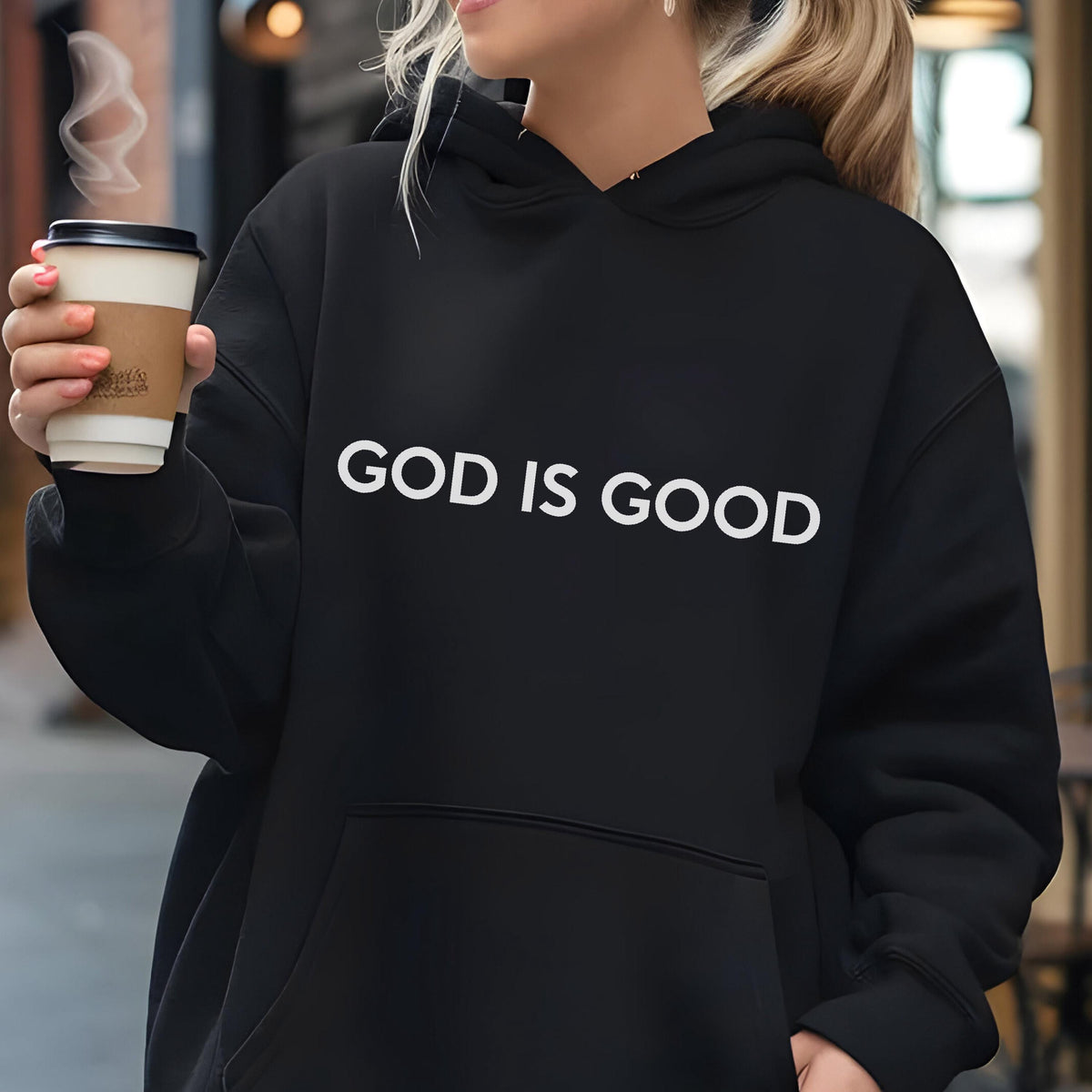 May include: Black hoodie with the white text "GOD IS GOOD". The hoodie has a drawstring hood and a kangaroo pocket. A person is holding a coffee cup with steam rising from it.