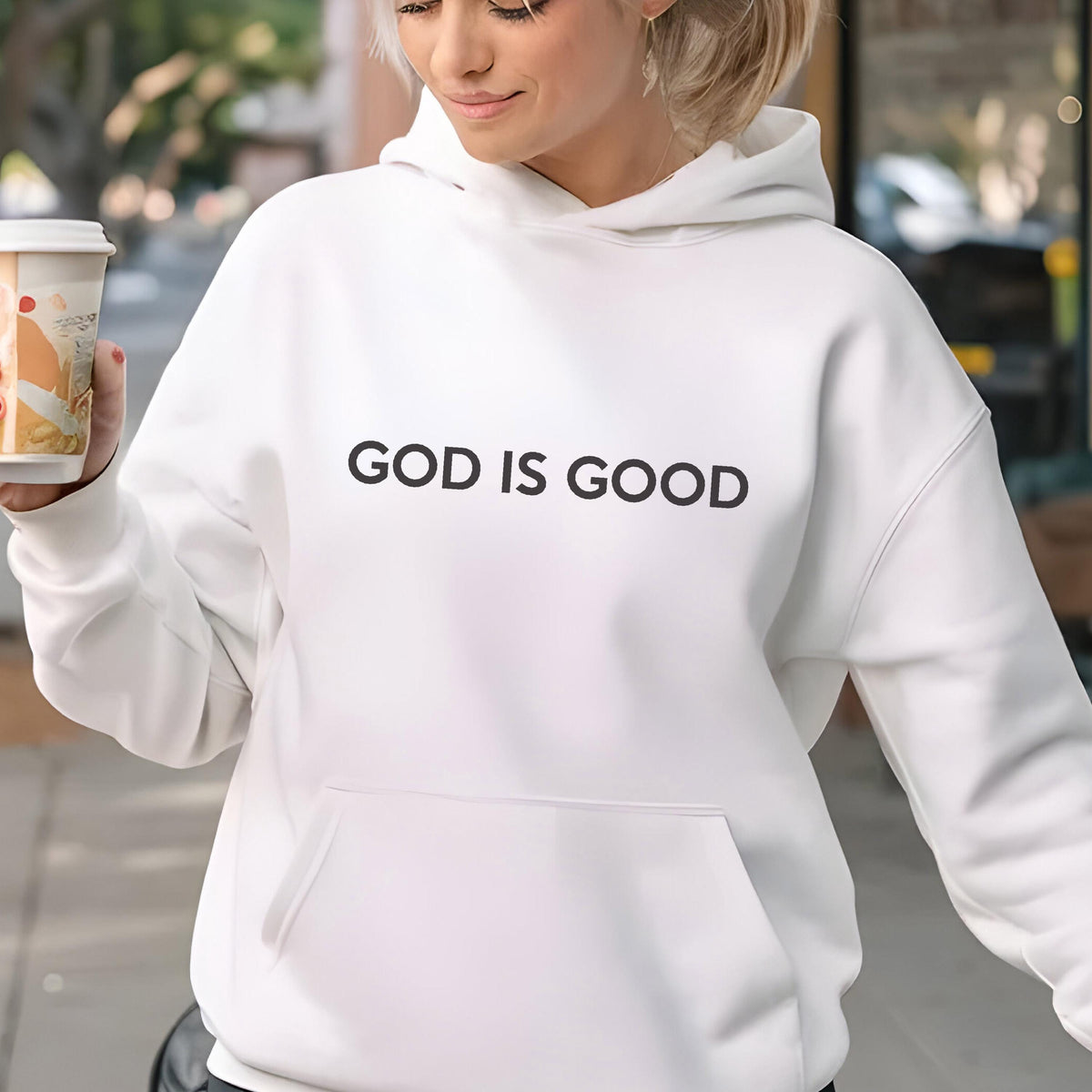 May include: White hoodie with the text "GOD IS GOOD" in black. The hoodie has a front pocket and a hood. The person in the image is holding a coffee cup.