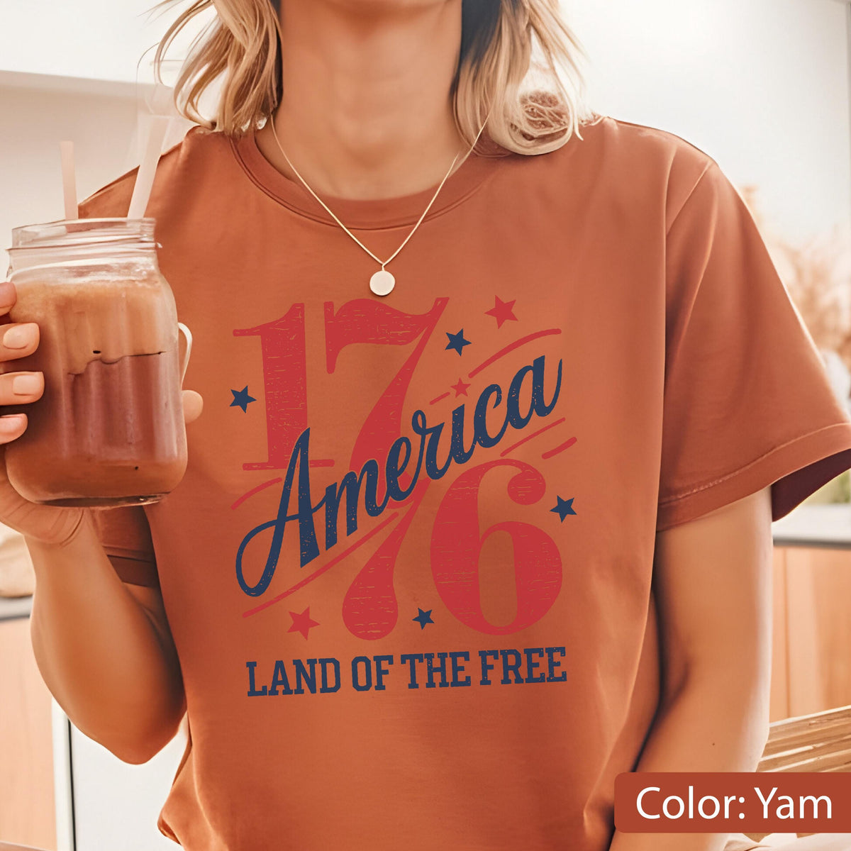 May include: A yam-colored t-shirt with a patriotic design. The design features the number "1776" in red, with the word "America" in blue script, and the phrase "Land of the Free" below. Small blue stars and red stars accent the design.