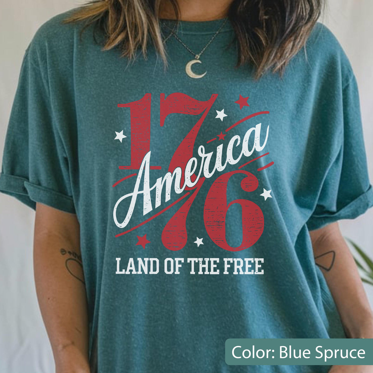 May include: Blue spruce t-shirt with the text "1776 America Land of the Free" in red and white. The design includes stars and the number 76 in a bold font. The shirt has rolled-up sleeves.