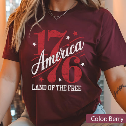 May include: Berry-colored t-shirt with the text "1776 America" in red and white, along with the phrase "LAND OF THE FREE." The design includes star accents. The shirt is a solid color.