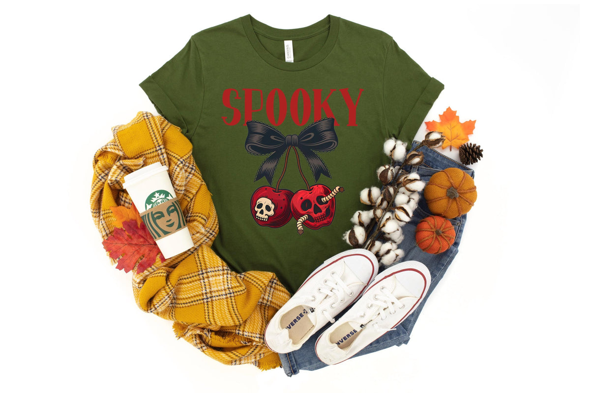 May include: An olive green t-shirt with the word "SPOOKY" in red, a black bow, and cherries with skull designs. The shirt is paired with jeans, white sneakers, and fall-themed decorations.