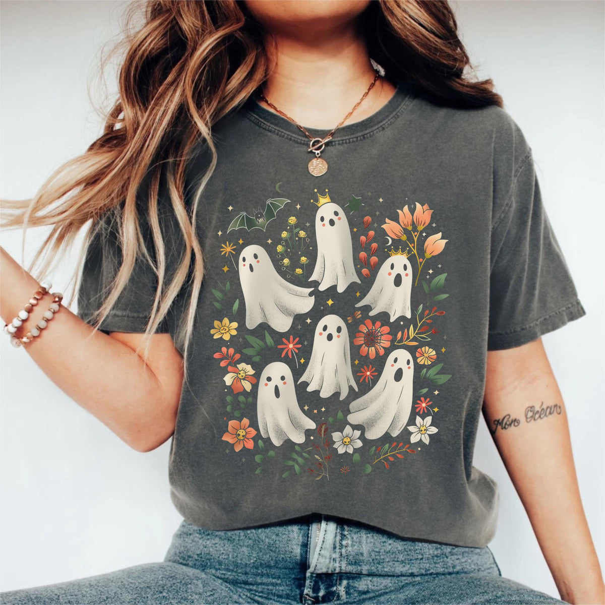 May include: Dark grey t-shirt featuring a whimsical design of cartoon ghosts surrounded by colorful flowers and foliage. The ghosts have simple faces and some wear crowns. A bat and crescent moon are also included in the design. The shirt is a comfortable, casual style.