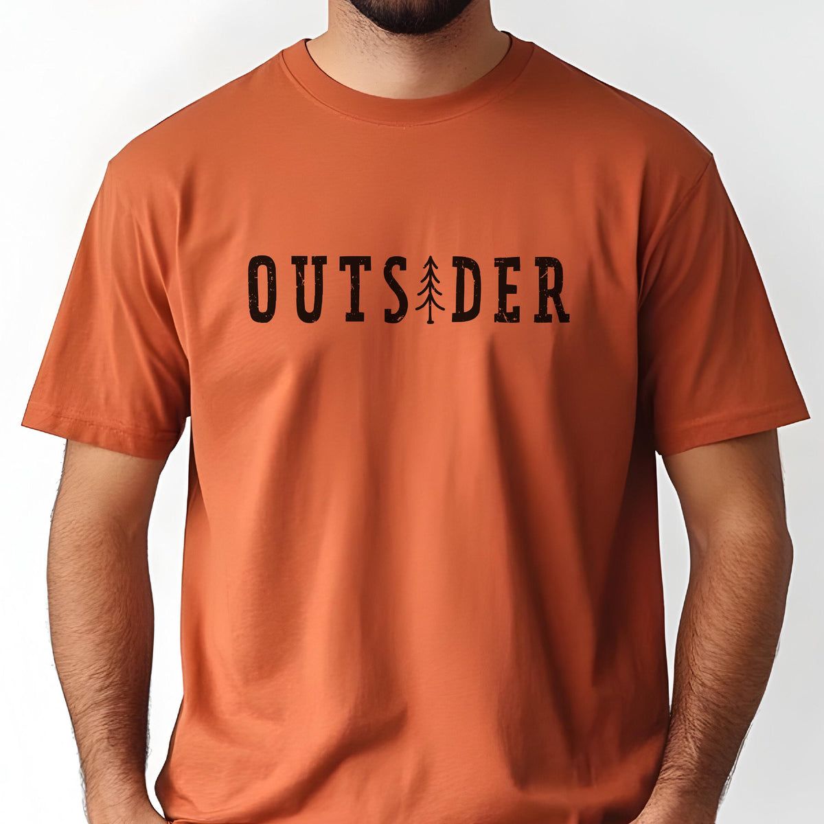 May include: A burnt orange short-sleeved t-shirt featuring the black text "OUTSIDER" printed across the chest. The letter "I" in the word is replaced by a stylized black graphic of a pine tree. The shirt has a crew neckline and a plain back.