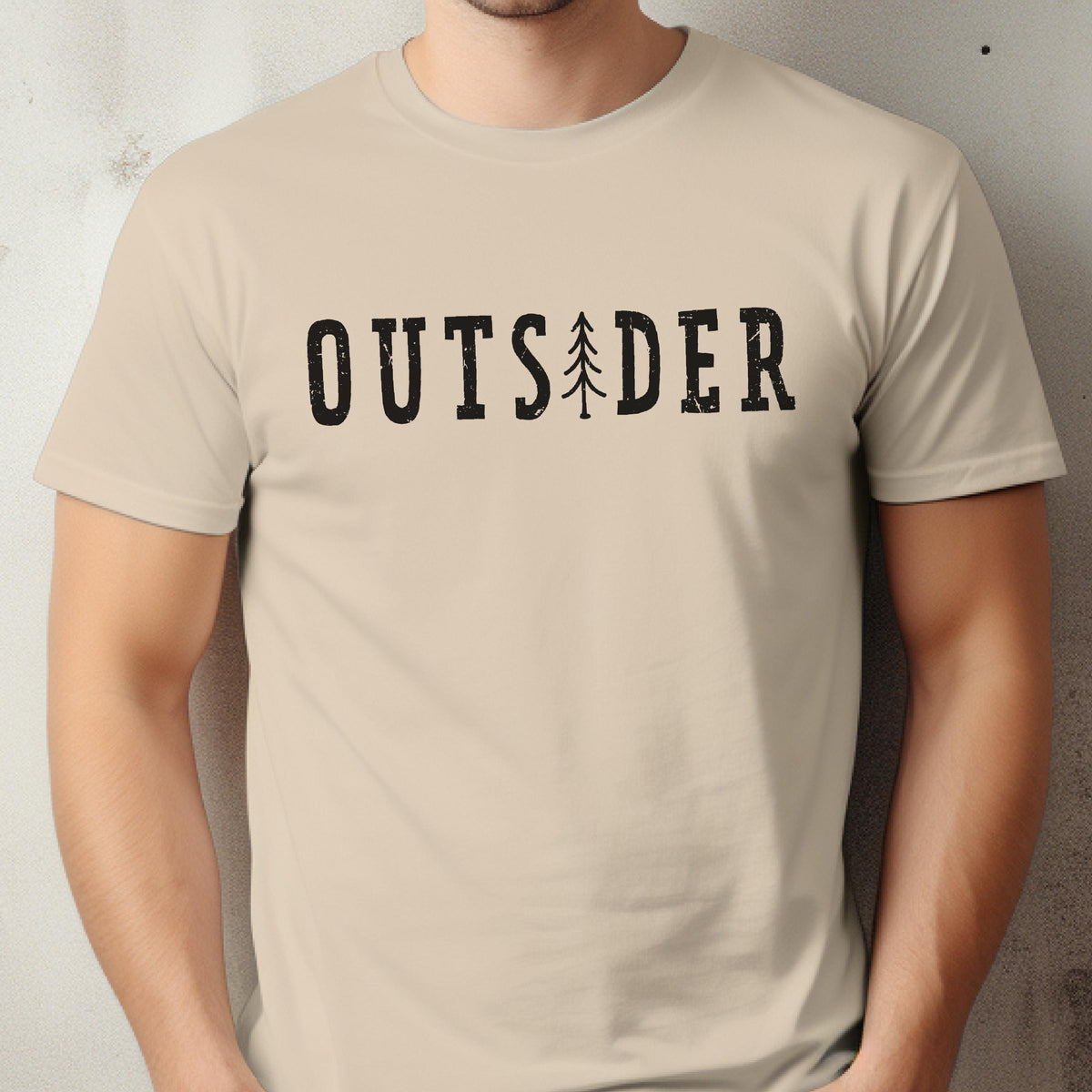 May include: A beige short-sleeved t-shirt featuring the black text "OUTSIDER" printed across the chest. The letter "I" in the word is replaced by a stylized black graphic of a pine tree. The font has a slightly distressed, textured appearance.