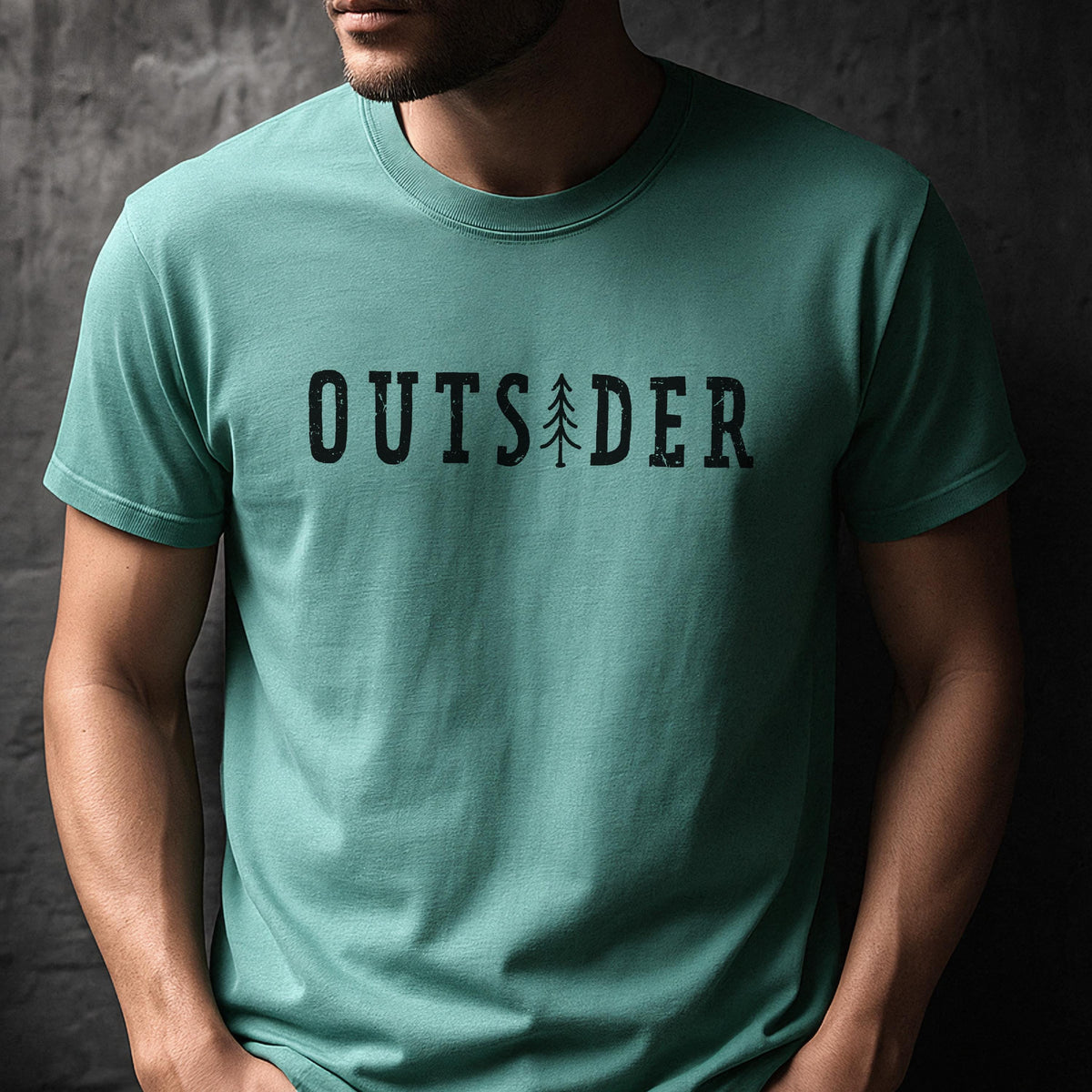 May include: A teal short-sleeved crew neck t-shirt featuring the word OUTSIDER printed in black across the chest. The letter I is replaced by a stylized black pine tree graphic. The text has a slightly distressed, weathered texture.