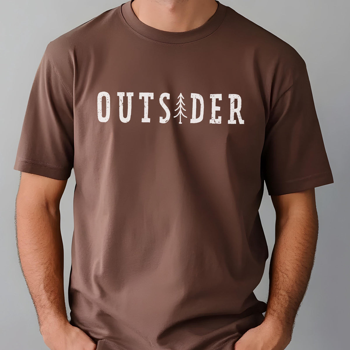 May include: A brown short-sleeved t-shirt featuring the white text OUTSIDER printed across the chest. The letter I is replaced by a stylized graphic of a pine tree. The font has a distressed, weathered texture throughout the lettering.