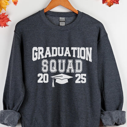 May include: Dark gray sweatshirt with the text "GRADUATION SQUAD 2025" in white block letters. A graduation cap graphic is between the numbers. The sweatshirt is displayed on a wooden hanger.