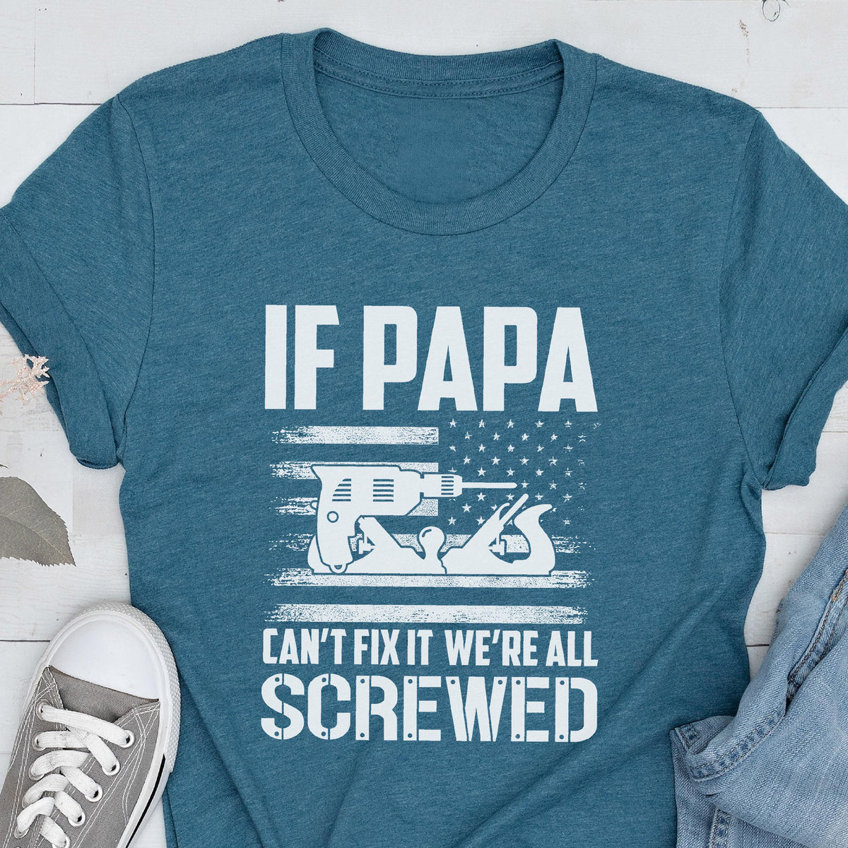 May include: Teal t-shirt with the text "IF PAPA CAN'T FIX IT WE'RE ALL SCREWED." The design includes a drill and a hand plane, set against a stylized American flag. A casual, humorous design.
