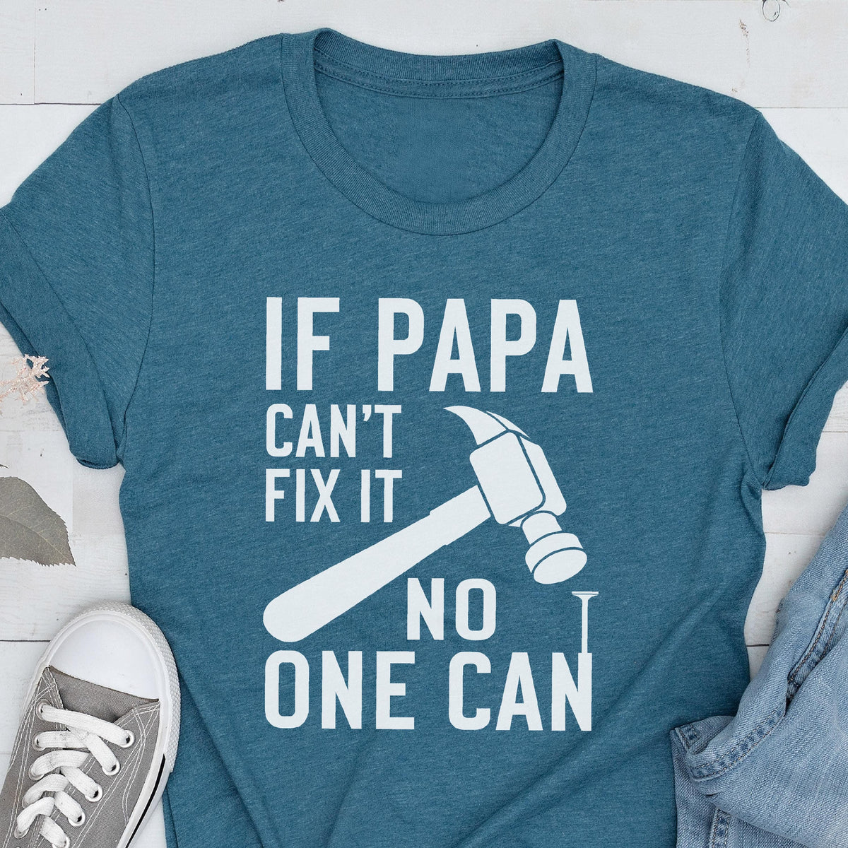 May include: Teal t-shirt with the text "IF PAPA CAN'T FIX IT NO ONE CAN" in white, along with a hammer and nail graphic. A gray sneaker and denim jeans are visible in the image.